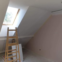 Dambud - Pokój na poddaszu w trakcie prac: ściana pomalowana na jasny róż, drewniana drabina oparta o ścianę pod oknem dachowym, widoczne ślady taśmy malarskiej przy suficie.