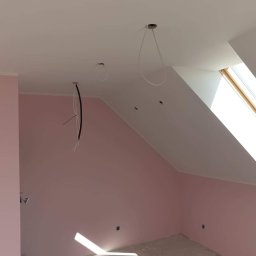 Dambud - Poddasze po malowaniu na różowo z widocznymi przewodami elektrycznymi wystającymi z sufitu, okno dachowe w skosie.