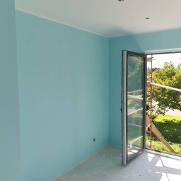 Dambud - Świeżo pomalowane na błękitno wnętrze z otwartym oknem balkonowym, przez które widać rusztowanie i zieleń ogrodu.