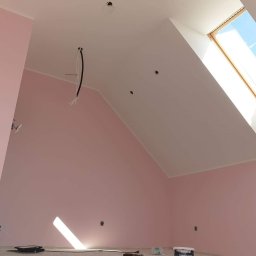 Dambud - Poddasze po malowaniu na różowo, z białym skośnym sufitem i oknem dachowym; widoczne przewody elektryczne, puszka z farbą na podłodze i narzędzia malarskie.