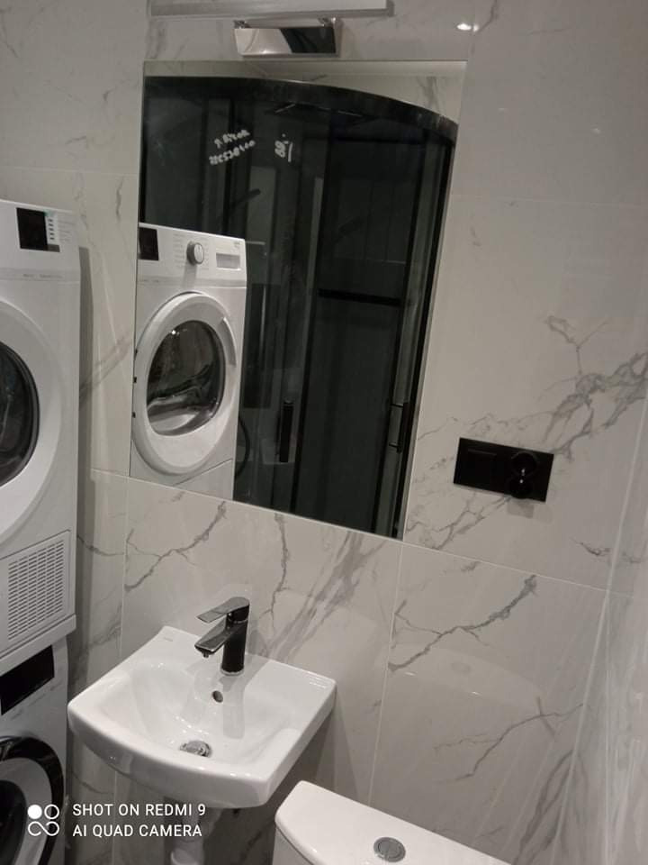 Mała łazienka w szarych płytkach imitujących marmur, z umywalką nablatową, toaletą, pralką i suszarką w zabudowie oraz lustrem, w którym odbija się kabina prysznicowa.