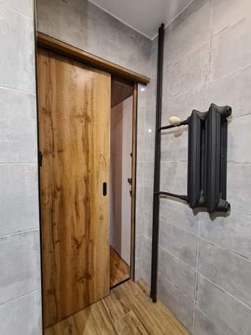 Wnętrze łazienki z przesuwnymi drzwiami z drewnopodobnym fornirem i grzejnikiem żeberkowym w kolorze czarnym, zamontowanym na ścianie wyłożonej szarymi płytkami.