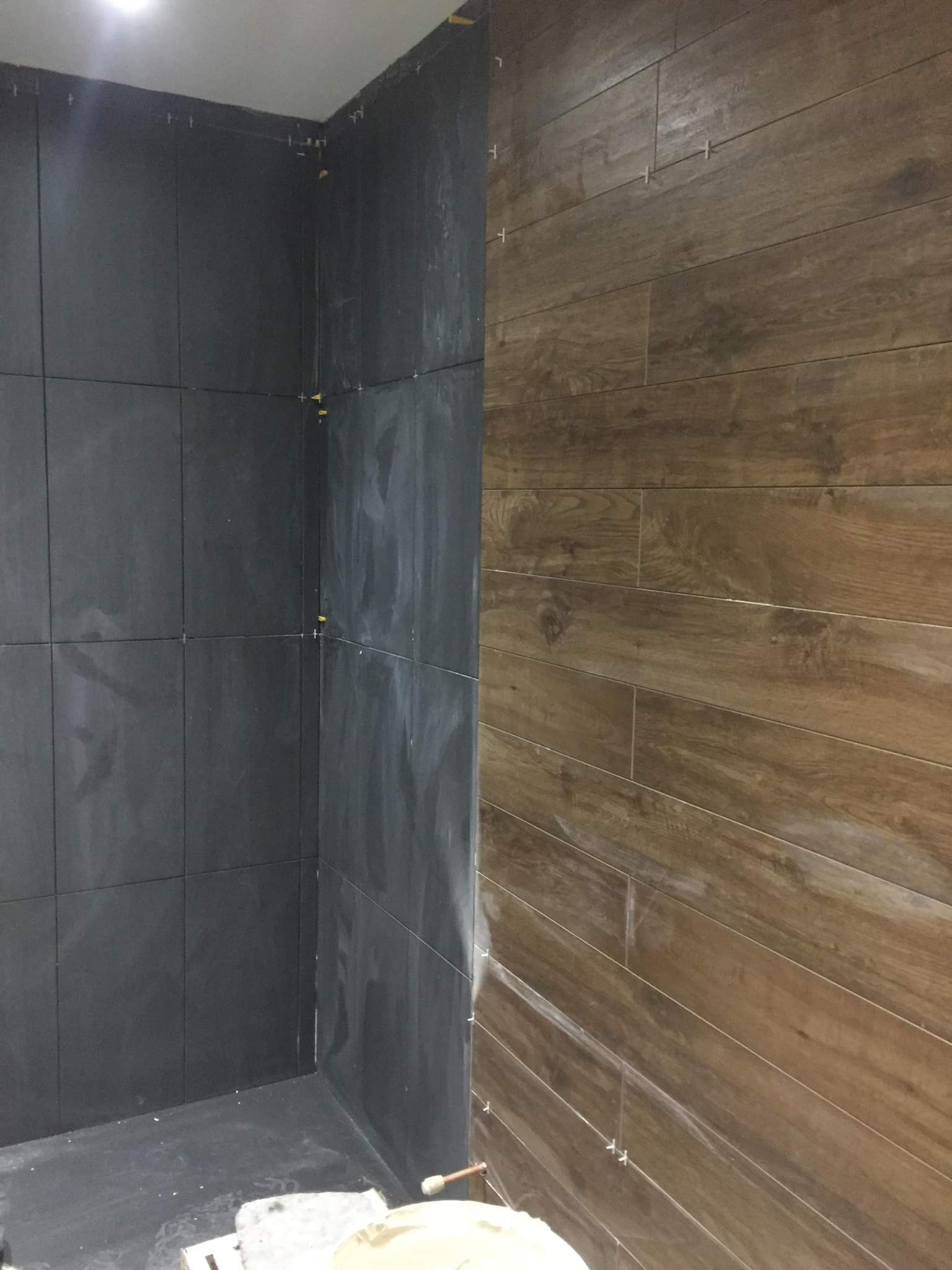 Układanie płytek imitujących drewno na ścianie obok ciemnych, kwadratowych płytek w rogu łazienki, widoczne krzyżyki dystansowe.