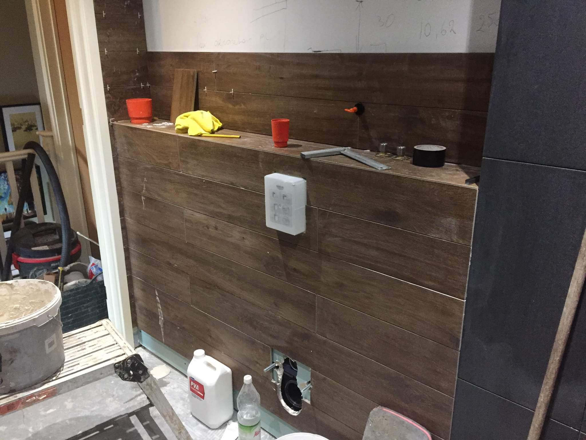 Montaż płytek imitujących drewno w łazience, widoczne narzędzia, poziomica, pojemniki, biała puszka z tworzywa sztucznego i otwór na instalację sanitarną.