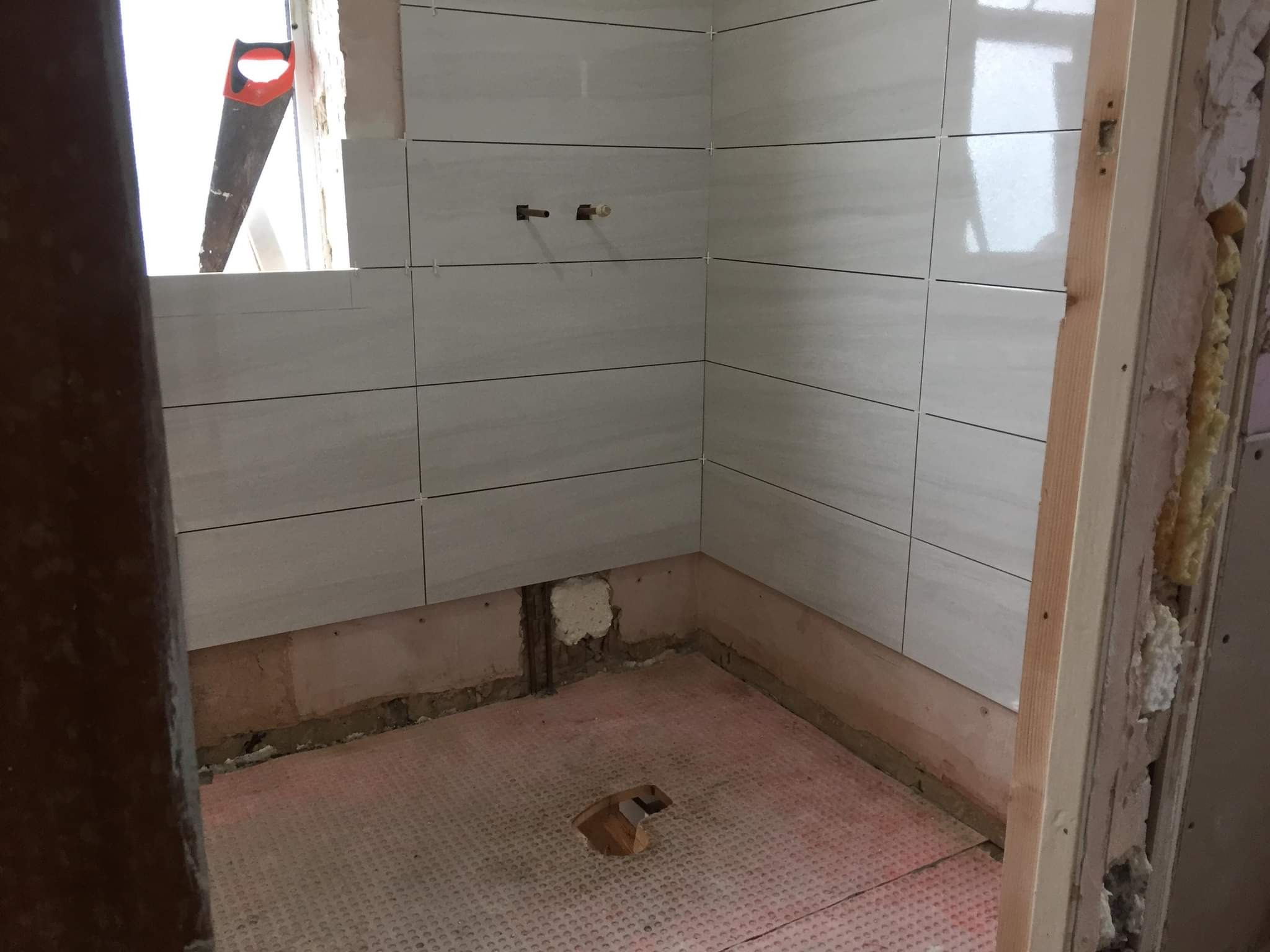 Łazienka w trakcie remontu: położone białe, poziome płytki na ścianach, widoczna piła ręczna, podłoga przygotowana pod płytki, odsłonięte instalacje, surowy stan wykończenia.