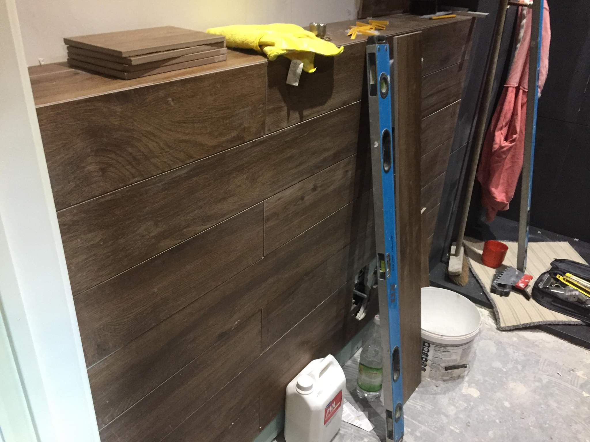 Montaż paneli ściennych imitujących drewno, widoczny poziomica, wiadro z klejem i zapas płytek, żółta szmatka na górnej krawędzi, surowy stan podłogi.