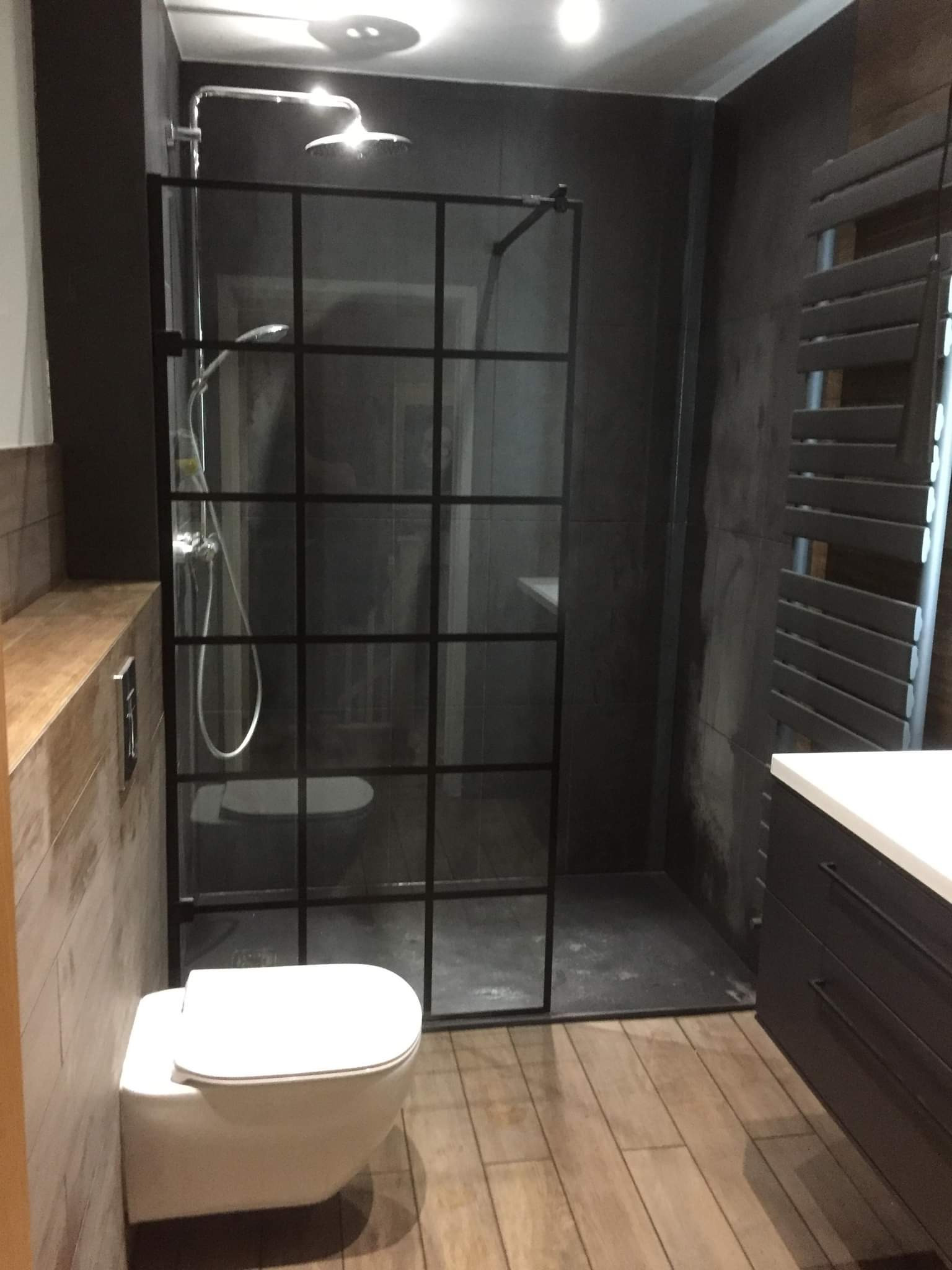 Nowoczesna łazienka z kabiną prysznicową typu walk-in z czarnymi profilami, drewnopodobnymi płytkami na podłodze i ścianie, oraz ciemnoszarą ścianą prysznicową.