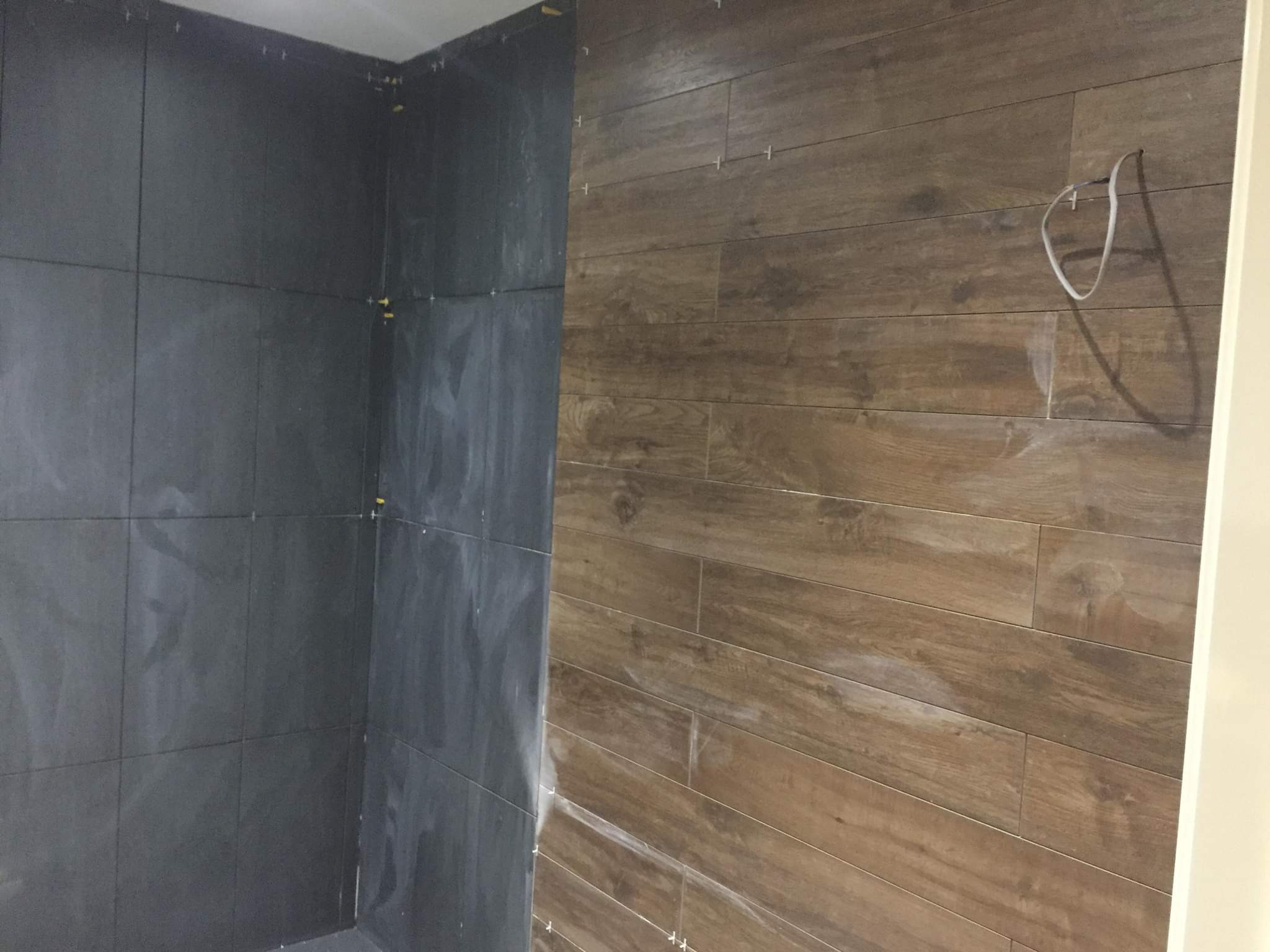 Ściana w trakcie wykańczania: Po lewej stronie ciemne, kwadratowe płytki, po prawej płytki imitujące drewno ułożone poziomo, widoczne krzyżyki dystansowe i wystający przewód elektryczny.