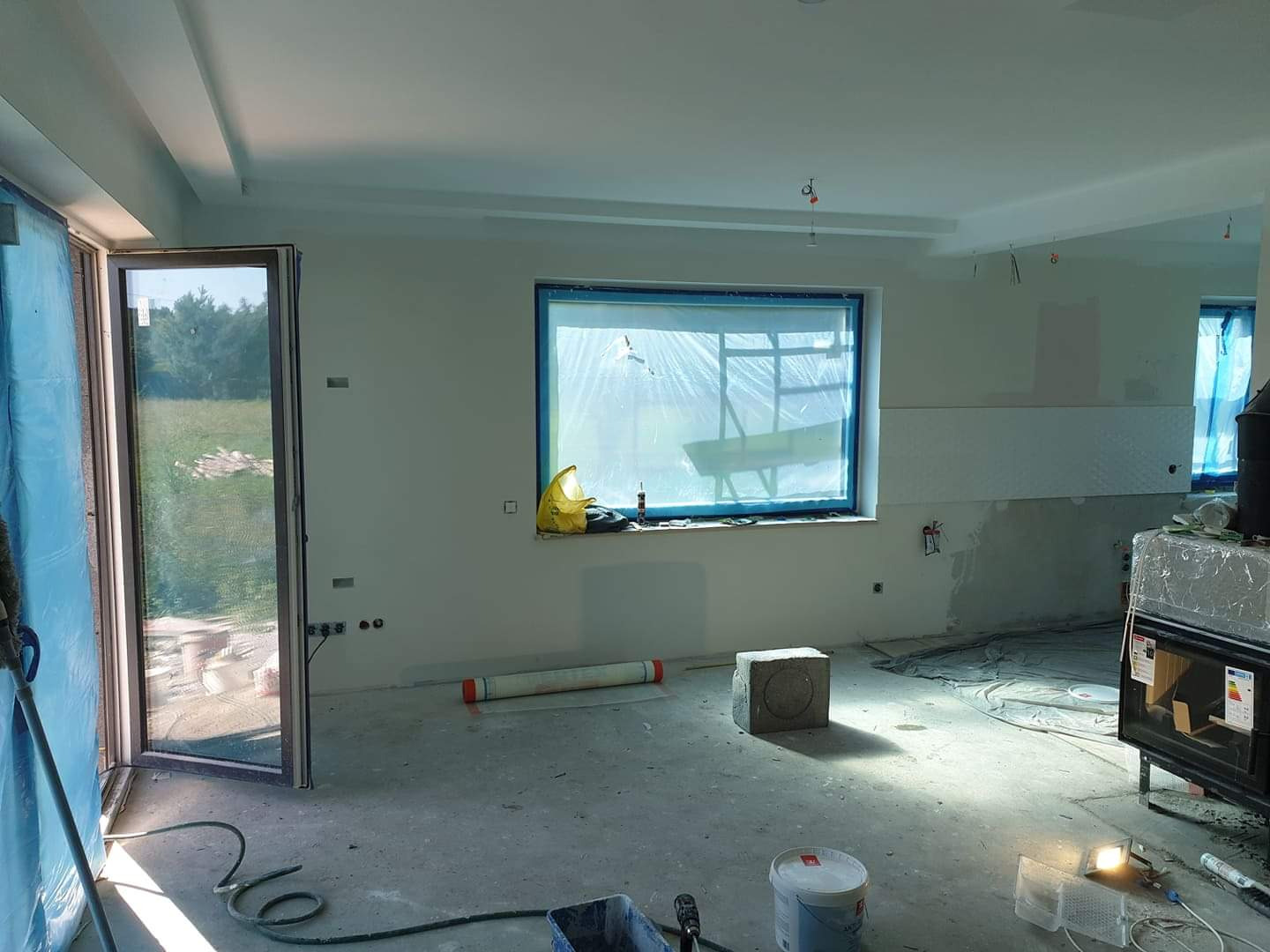 Wnętrze w trakcie prac wykończeniowych: zabezpieczone okna folią, świeżo pomalowane ściany, surowa podłoga, widoczne narzędzia i materiały budowlane, otwarte drzwi balkonowe z widokiem na zieleń.
