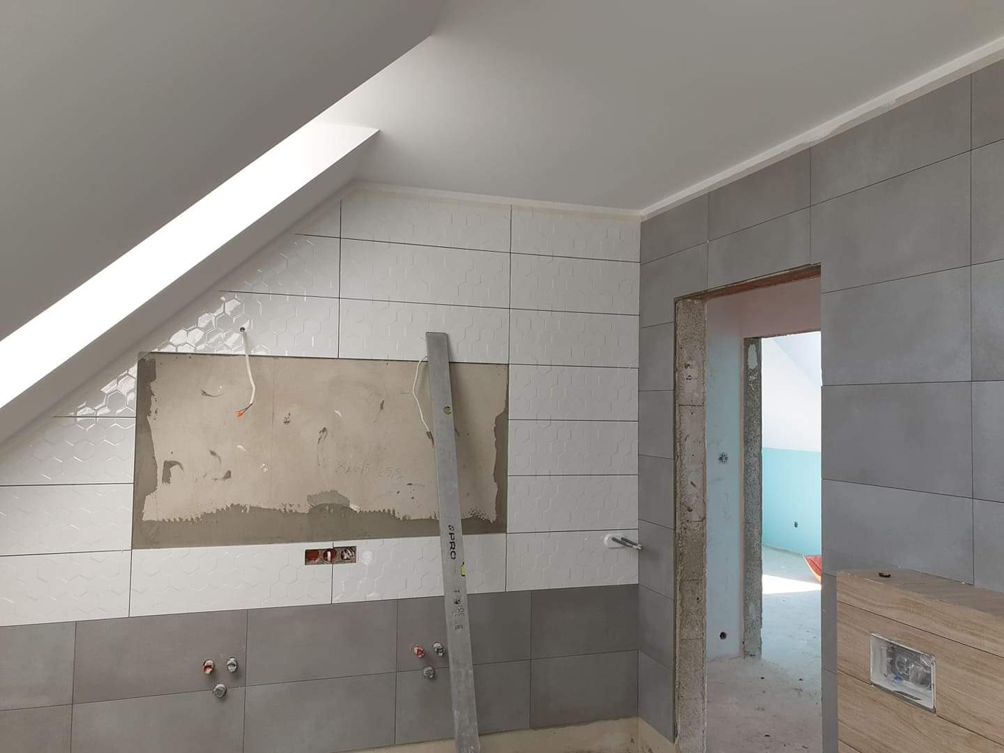 Układanie szarych i białych płytek heksagonalnych w łazience na poddaszu, widoczny fragment ściany bez płytek z wystającymi kablami i poziomica oparta o ścianę.