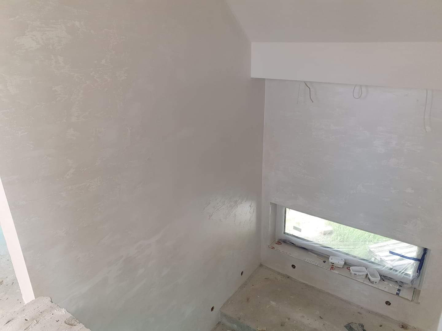 Ściany pokryte dekoracyjną, jasnoszarą strukturą w nowym budynku, z widocznymi puszkami elektrycznymi w ścianie i zabezpieczonym oknem.