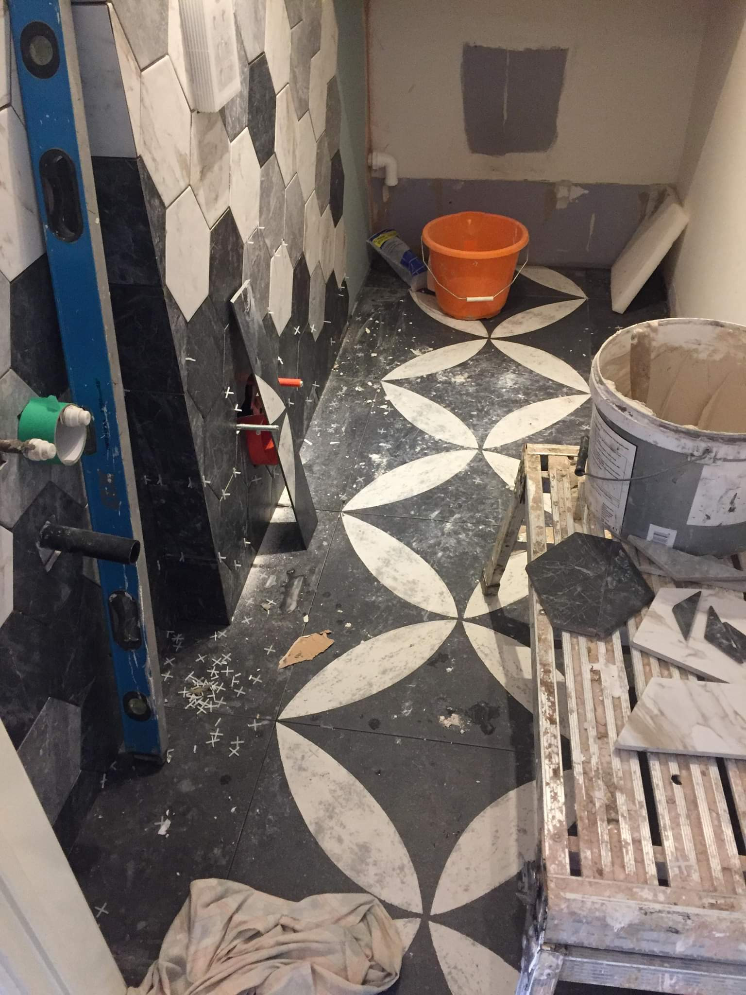 Układanie heksagonalnych płytek podłogowych i ściennych w łazience, widoczny poziomica, wiadro, krzyżyki dystansowe i drabina, praca w toku.