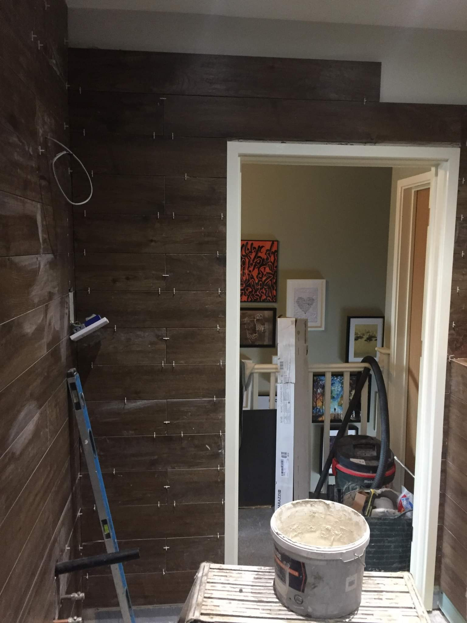 Montaż brązowych płytek imitujących drewno na ścianie w trakcie prac, widoczna poziomica i wiadro z zaprawą, w tle korytarz z obrazami.