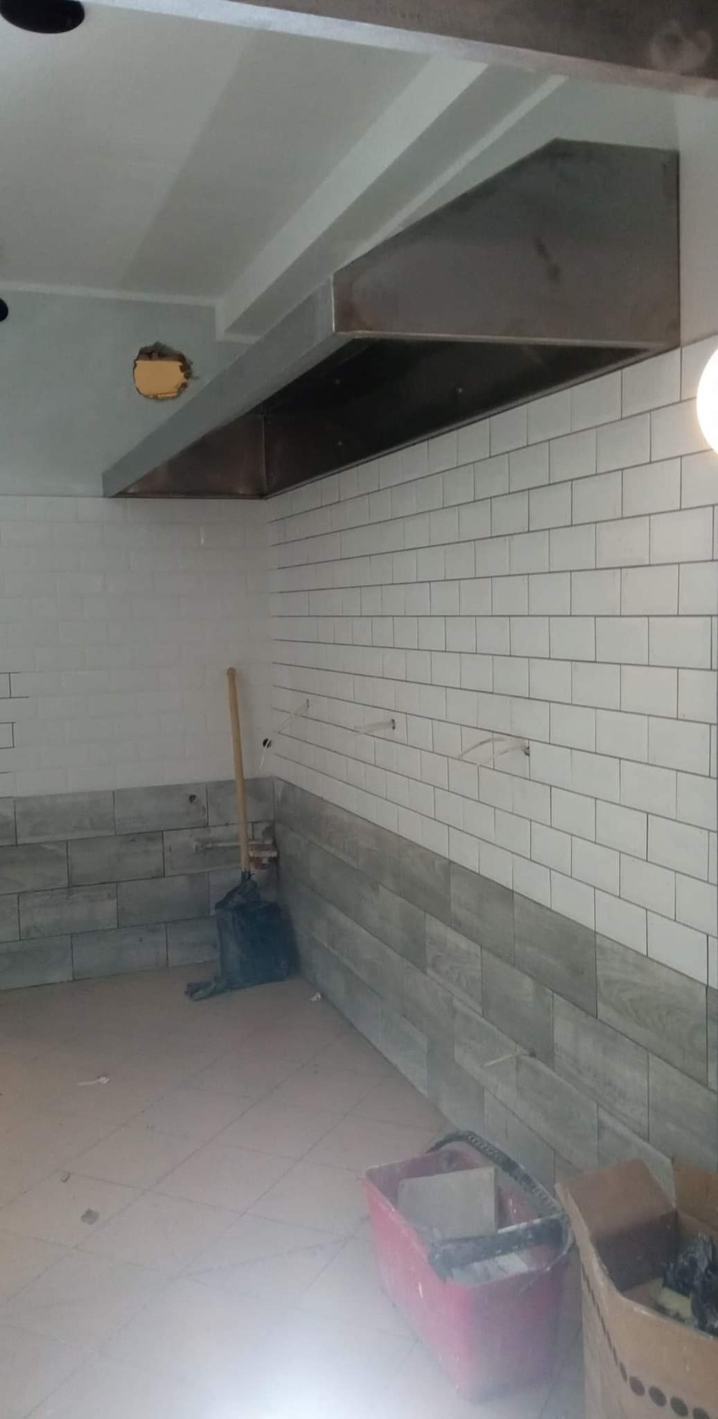 Wnętrze lokalu gastronomicznego w trakcie prac wykończeniowych: białe płytki ścienne, szary pas płytek imitujących drewno u dołu, zamontowany okap ze stali nierdzewnej, widoczne narzędzia...