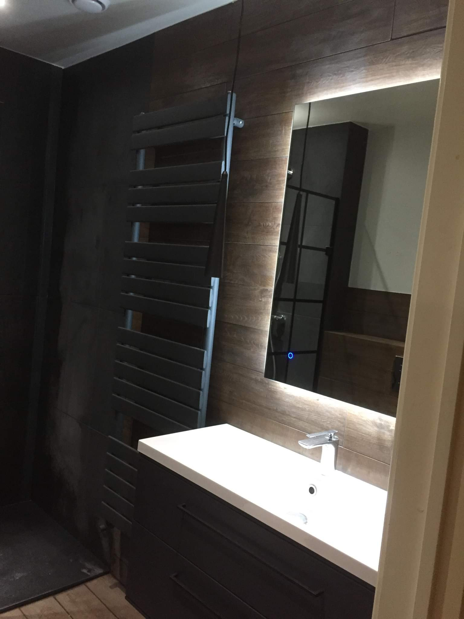 Nowoczesna łazienka z ciemnymi płytkami imitującymi drewno, grzejnikiem drabinkowym i podświetlanym lustrem nad białą umywalką z szafką.