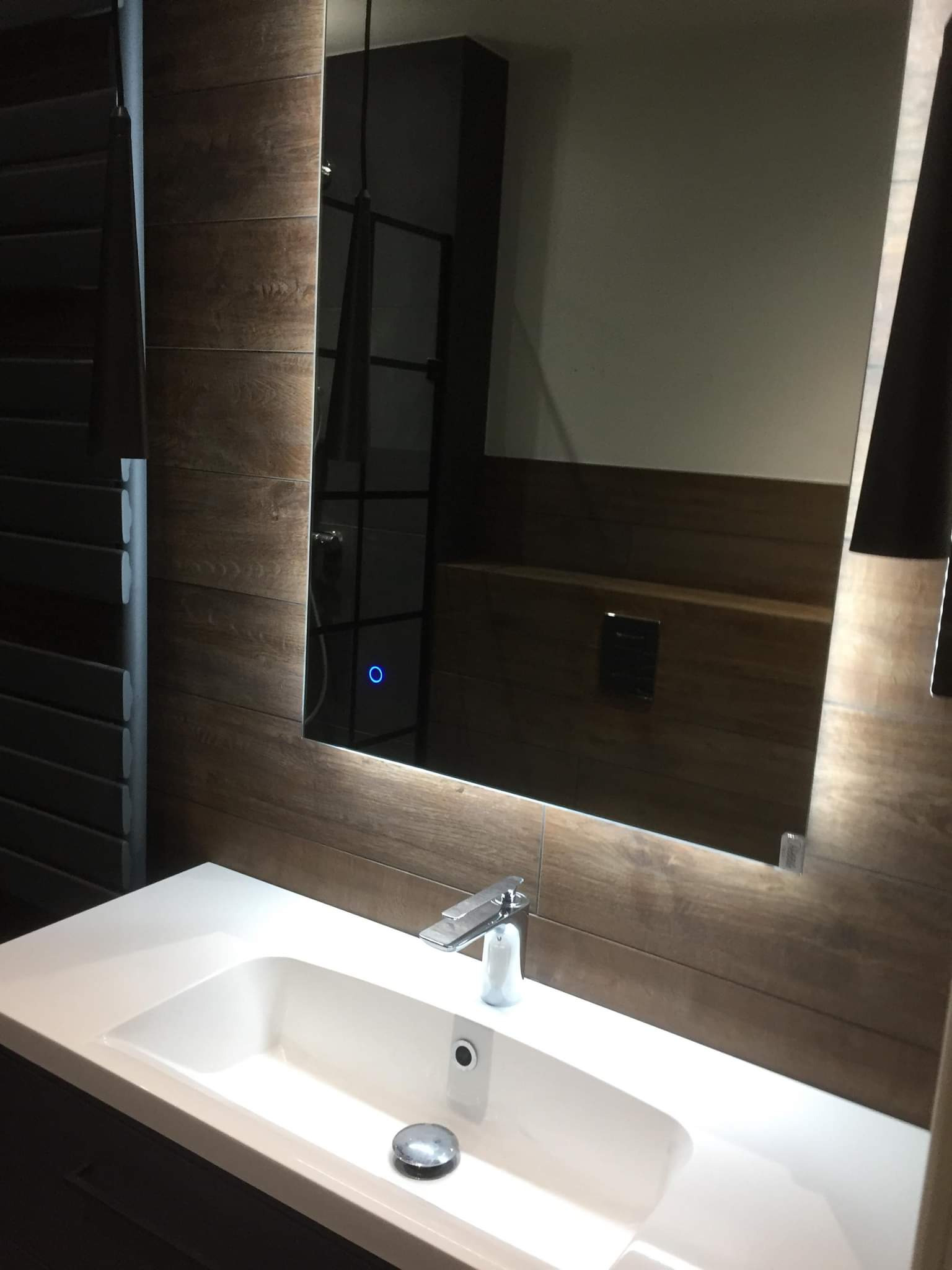 Nowoczesna łazienka z umywalką wpuszczaną w blat, podświetlanym lustrem z wbudowanym włącznikiem dotykowym i drewnopodobnymi płytkami na ścianie.