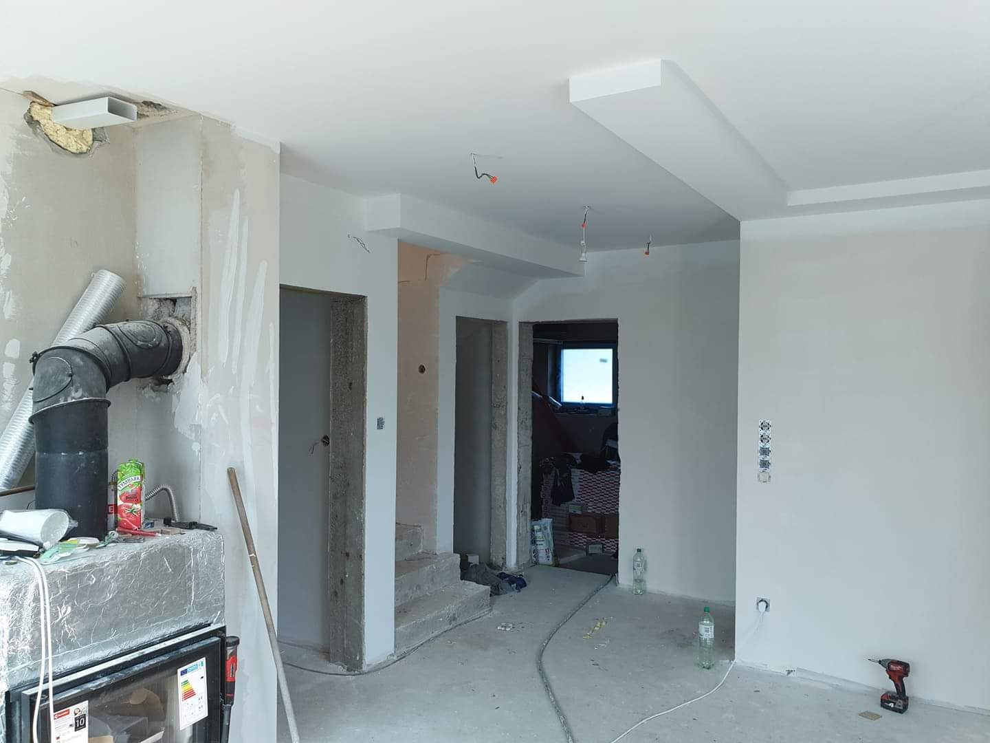 Wnętrze w trakcie prac, widoczne surowe ściany, instalacja kominkowa z czarnymi rurami, częściowo wykończony sufit z podwieszanymi elementami, otwory drzwiowe bez ościeżnic, oraz narzędzia...