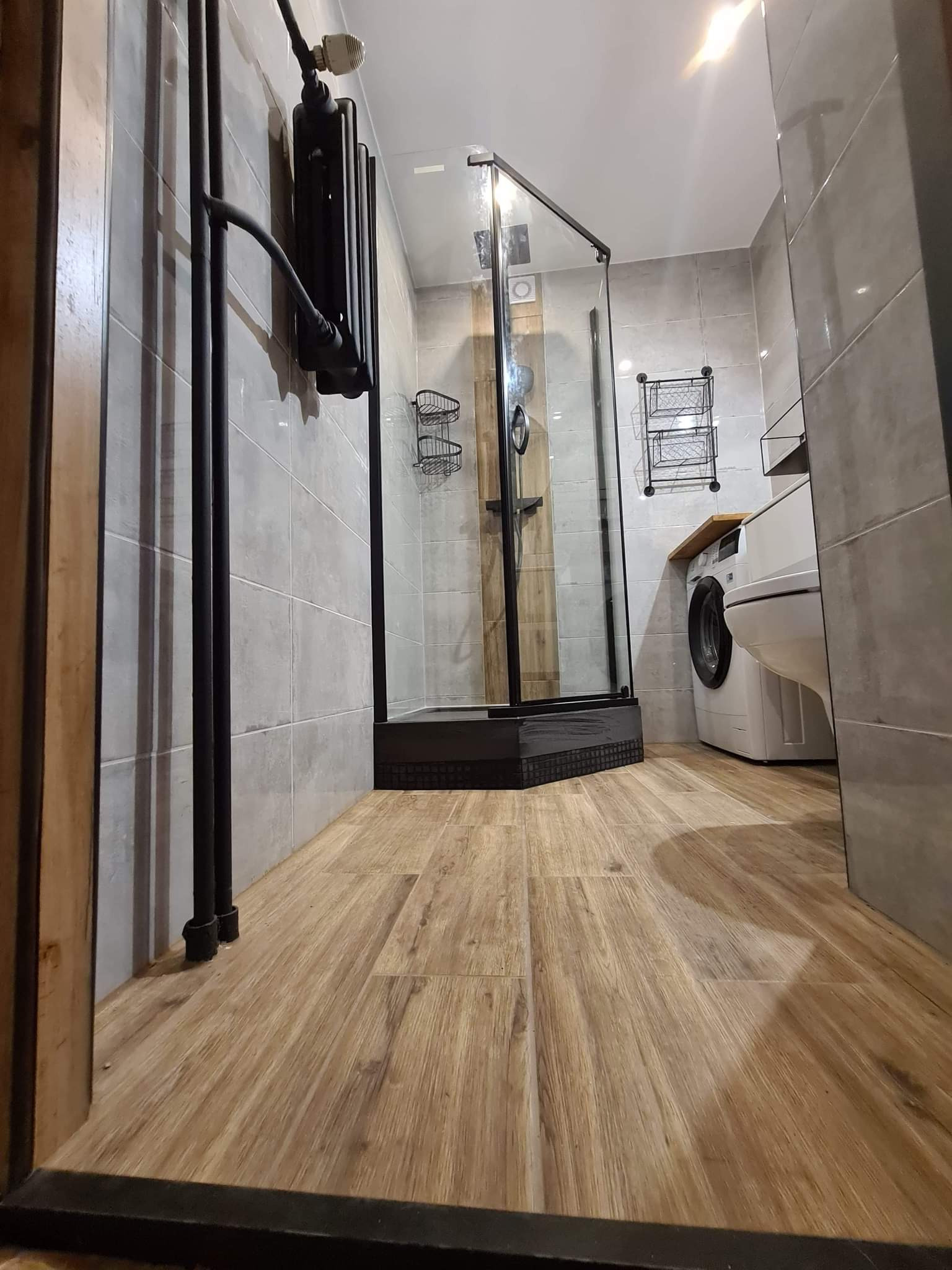 Nowoczesna łazienka z kabiną prysznicową typu walk-in z czarnymi profilami, grzejnikiem drabinkowym w stylu industrialnym, płytkami imitującymi beton i drewnianą podłogą laminowaną.