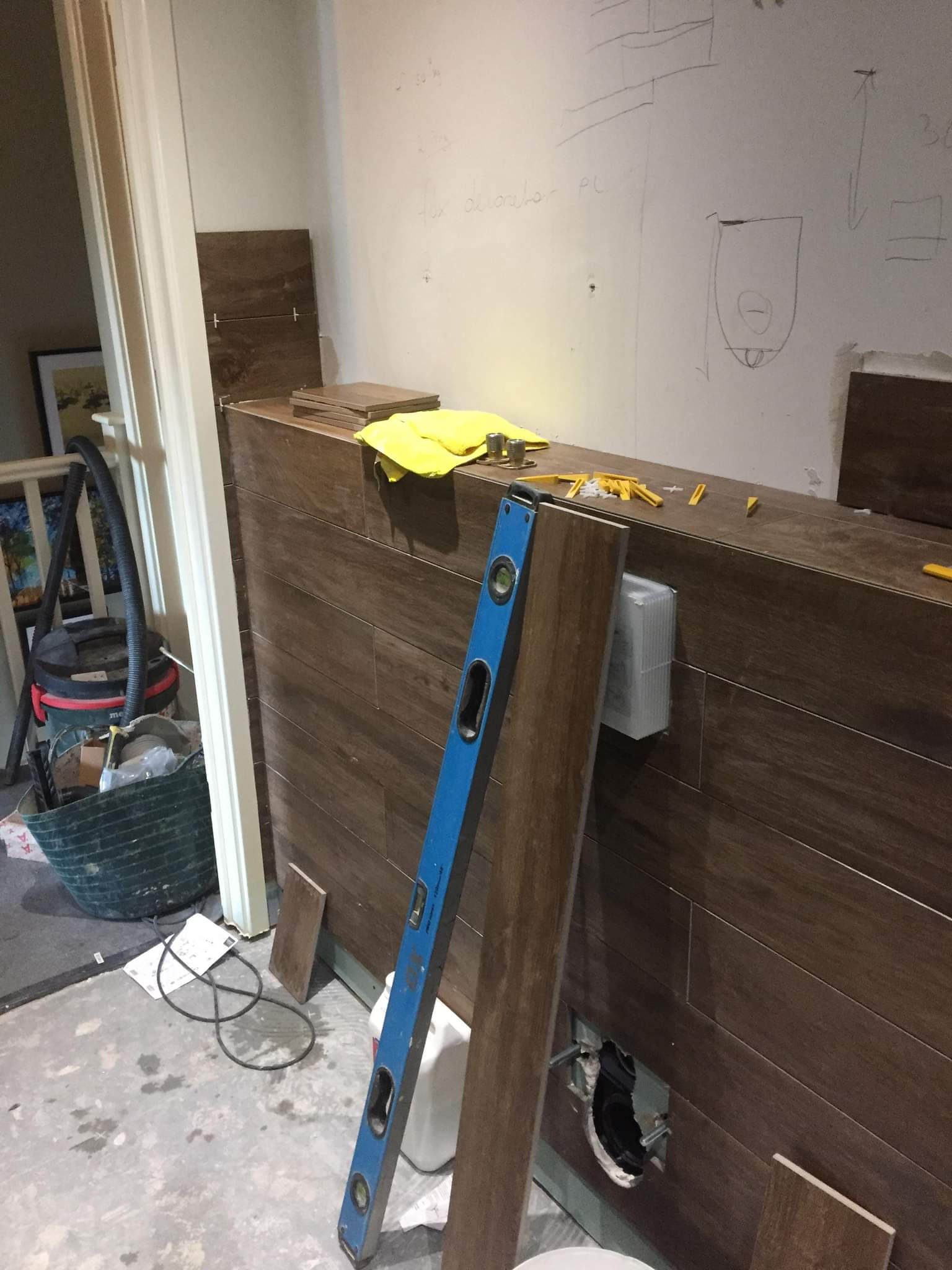 Montaż płytek imitujących drewno na ścianie, widoczna poziomica, zapas płytek, żółta ściereczka i akcesoria montażowe, na ścianie szkic instalacji.