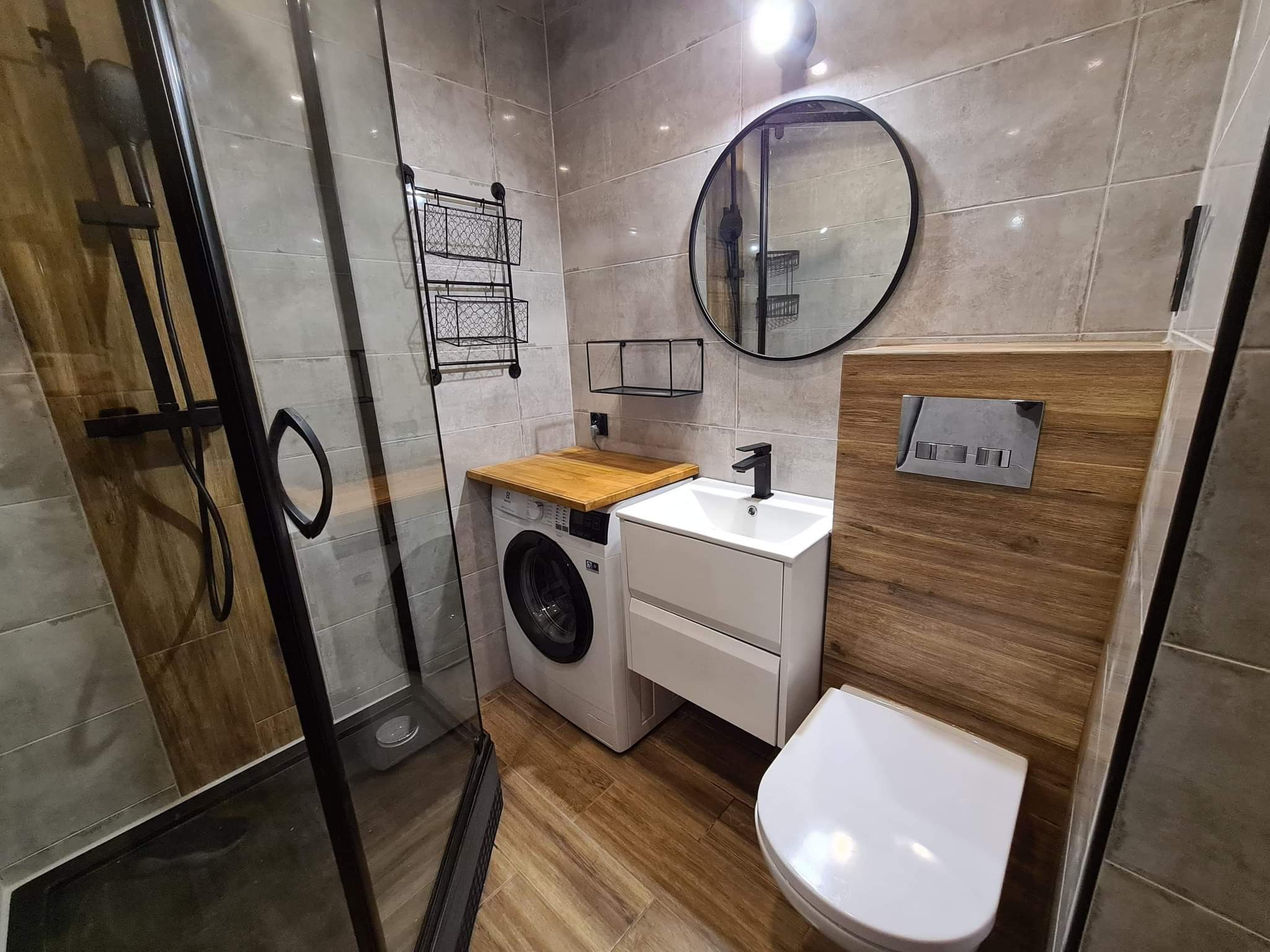 Nowoczesna łazienka z kabiną prysznicową typu walk-in, pralką z drewnianym blatem, umywalką z szafką i okrągłym lustrem w czarnej ramie, wykończona płytkami w odcieniach szarości i drewna.