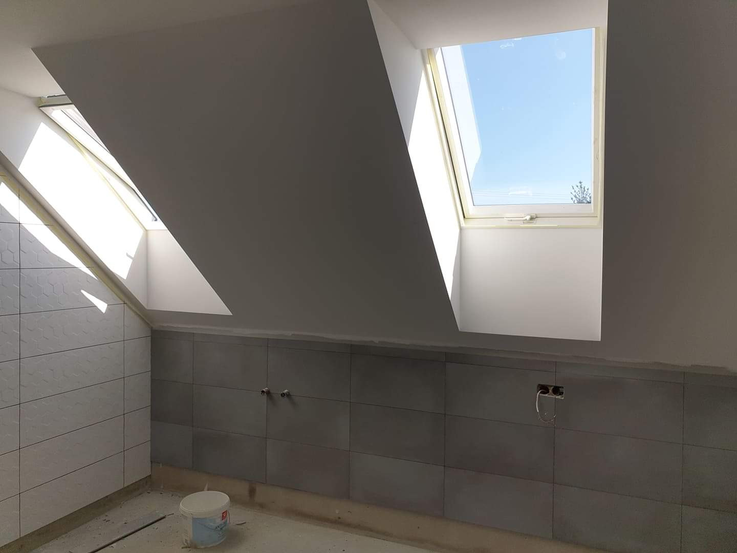 Poddasze w trakcie wykańczania: szare płytki częściowo ułożone na ścianie, dwa okna dachowe wpuszczające jasne światło, widoczne puszki elektryczne bez osprzętu, wiaderko z farbą na podłodze.