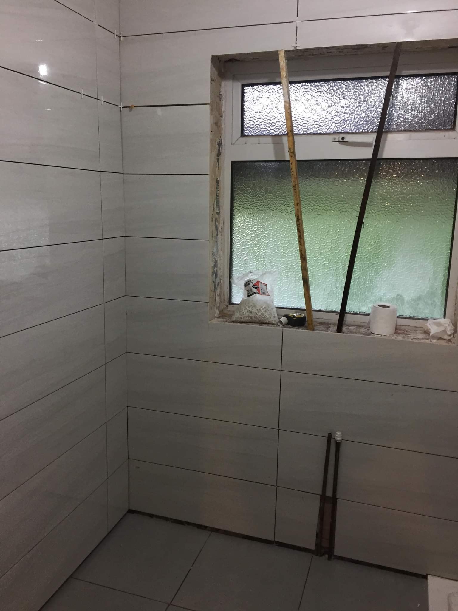 Świeżo wykafelkowana łazienka z szarymi płytkami, widoczne krzyżyki dystansowe, okno z szybą ornamentową, paczka z krzyżykami, poziomica, rolka papieru toaletowego i ślady po remoncie.