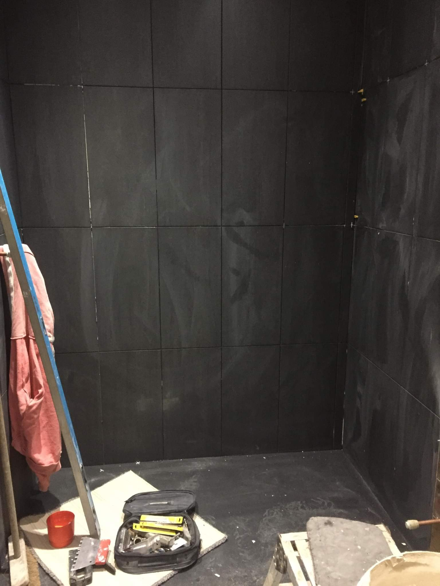 Montaż ciemnych, kwadratowych płytek na ścianie w rogu pomieszczenia, widoczne krzyżyki dystansowe, narzędzia w torbie, drabina oparta o ścianę.