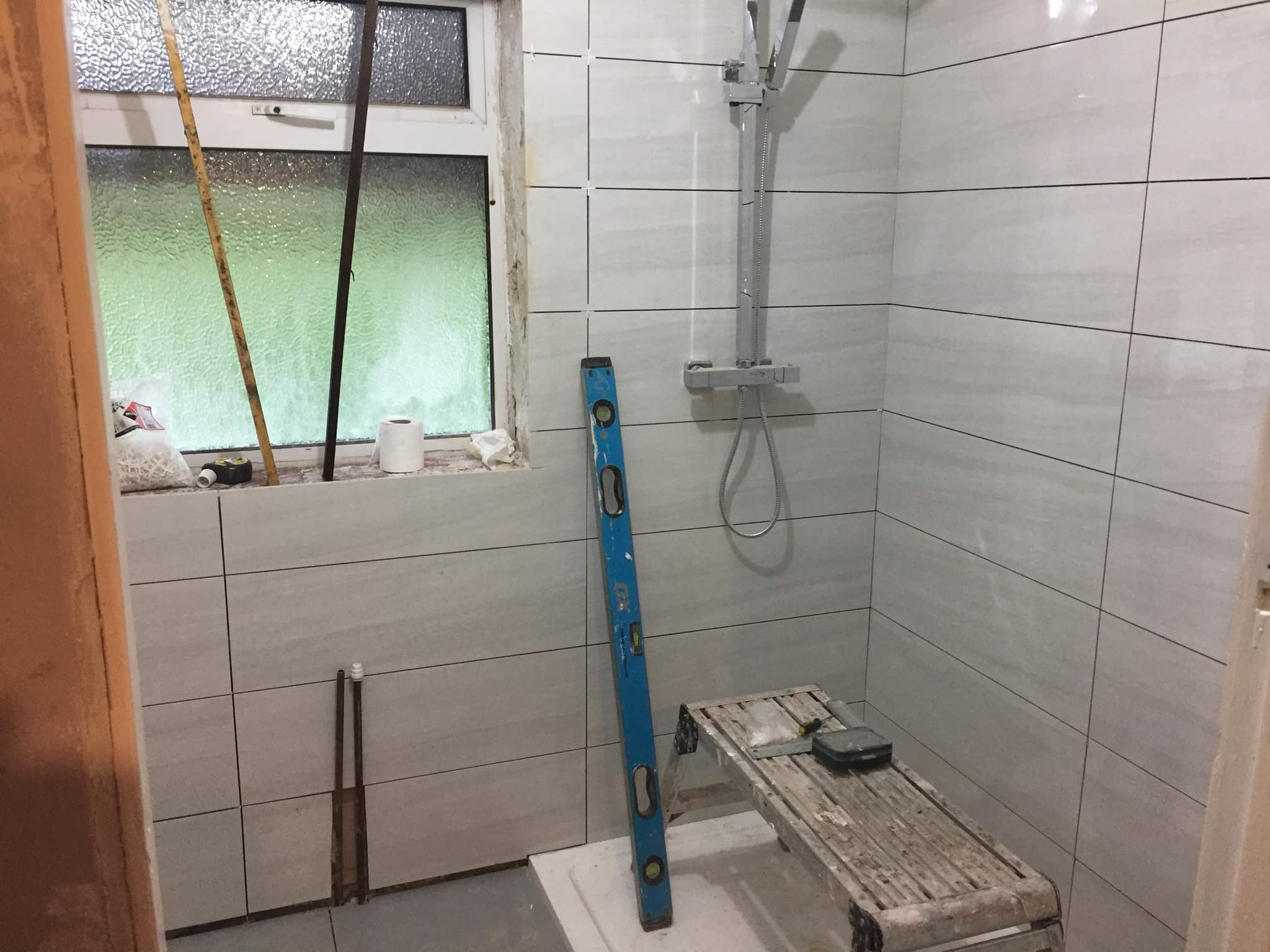 Łazienka w trakcie układania płytek ściennych, widoczna drabina malarska, poziomica i okno z widokiem na zieleń.