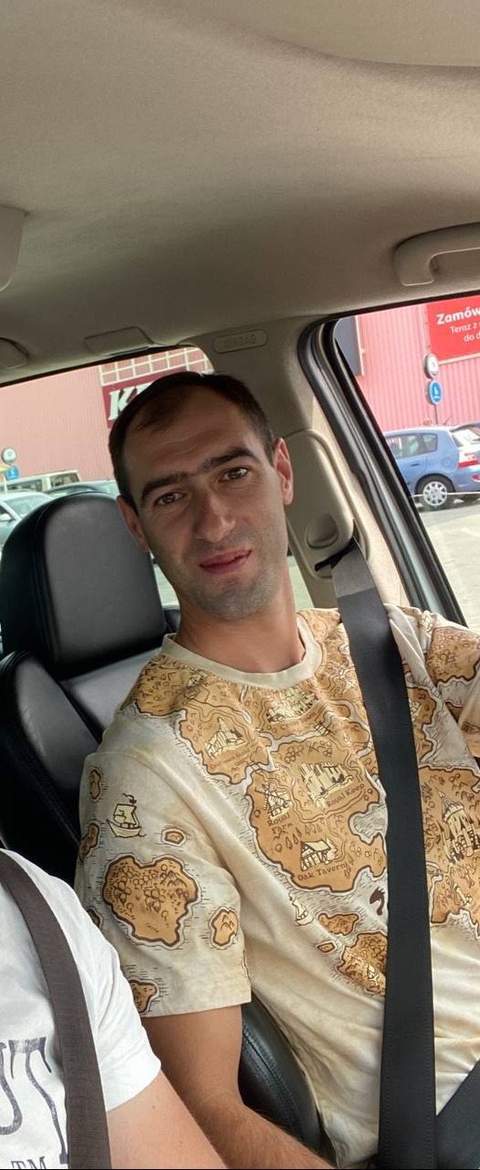 Mężczyzna w samochodzie, ubrany w koszulkę z motywem mapy, zapięty pasem bezpieczeństwa, na tle parkingu przed budynkiem sklepu budowlanego.