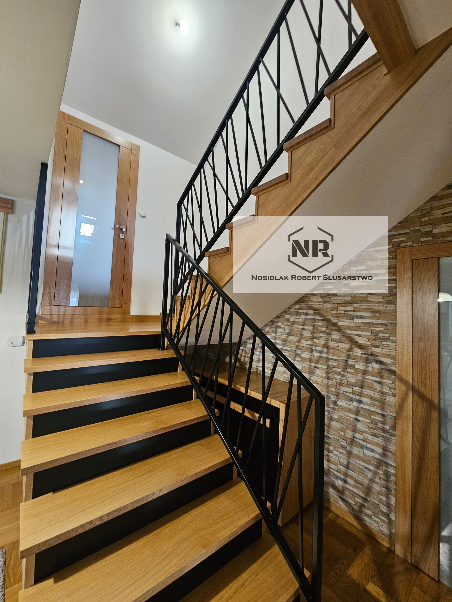 Drewniane schody z czarnymi podstopnicami i nowoczesną metalową balustradą o geometrycznym wzorze, widok z dołu na górę, ściana wykończona dekoracyjną okładziną imitującą kamień.