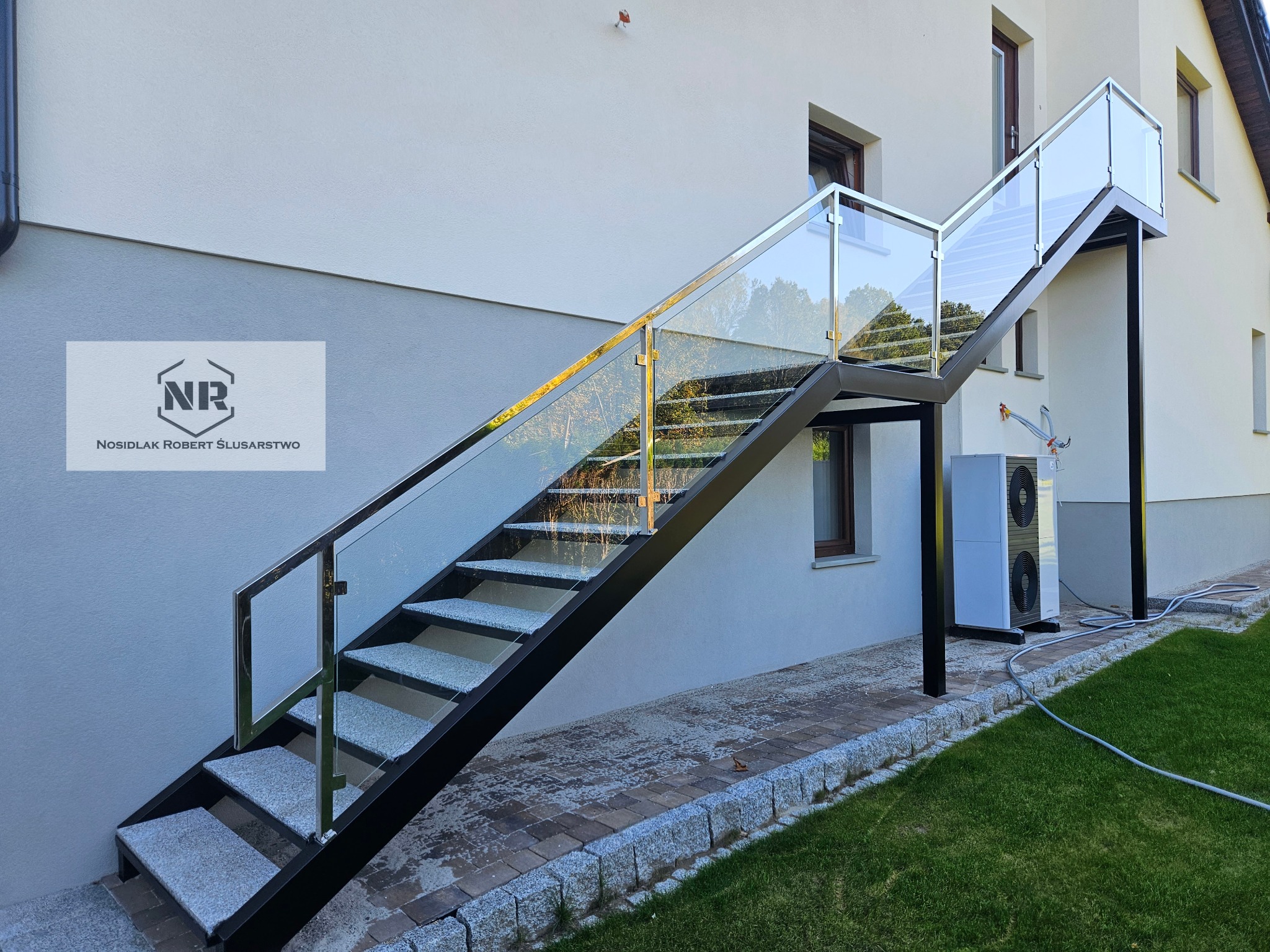 Zewnętrzne schody metalowe z szarymi stopniami i szklanymi balustradami, prowadzące do wejścia na piętro budynku, z widocznym logo firmy na ścianie.