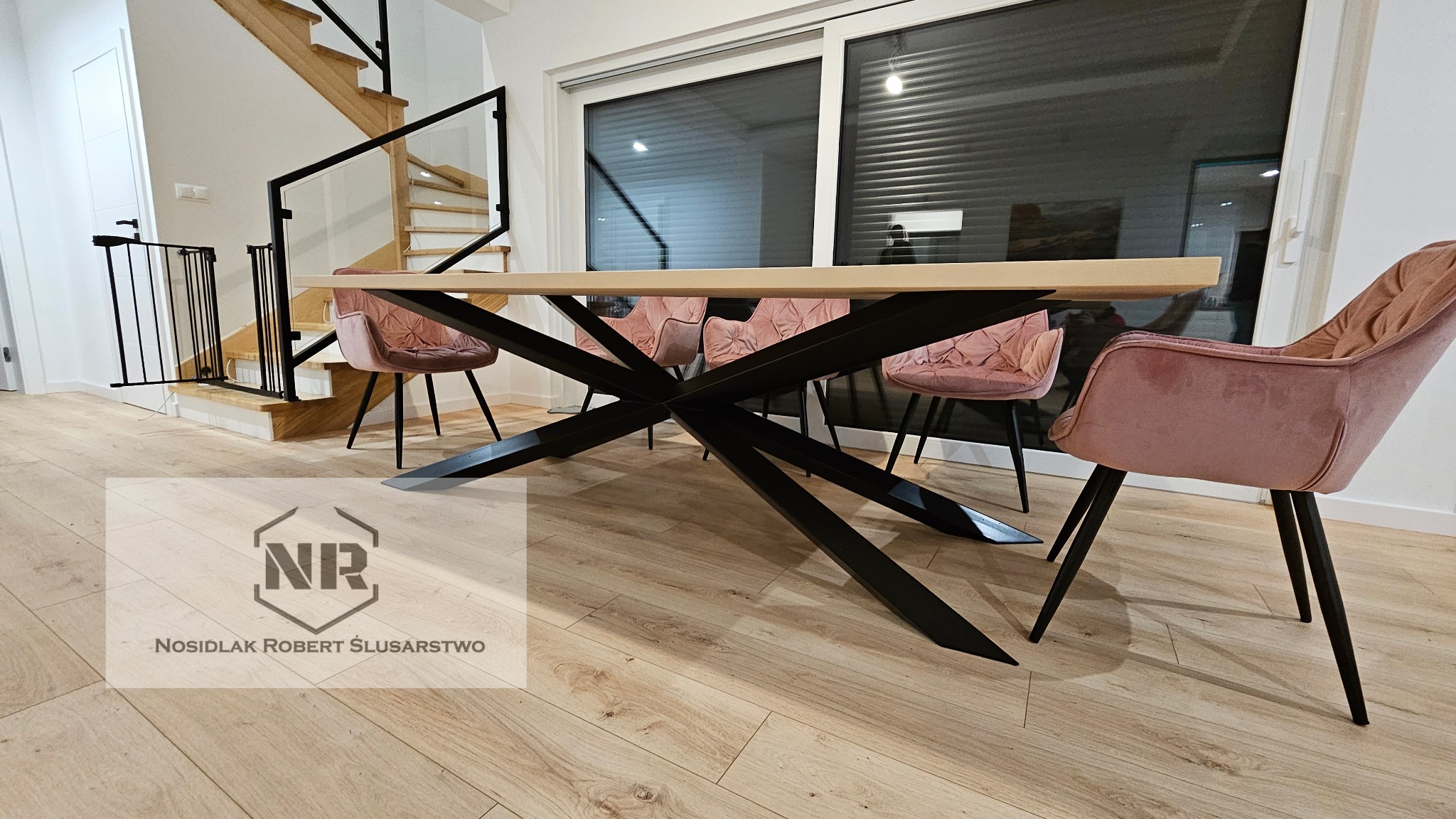Stół z jasnym blatem i metalową podstawą o geometrycznym kształcie, otoczony różowymi krzesłami na tle nowoczesnego wnętrza z drewnianymi schodami i oknem z żaluzjami.