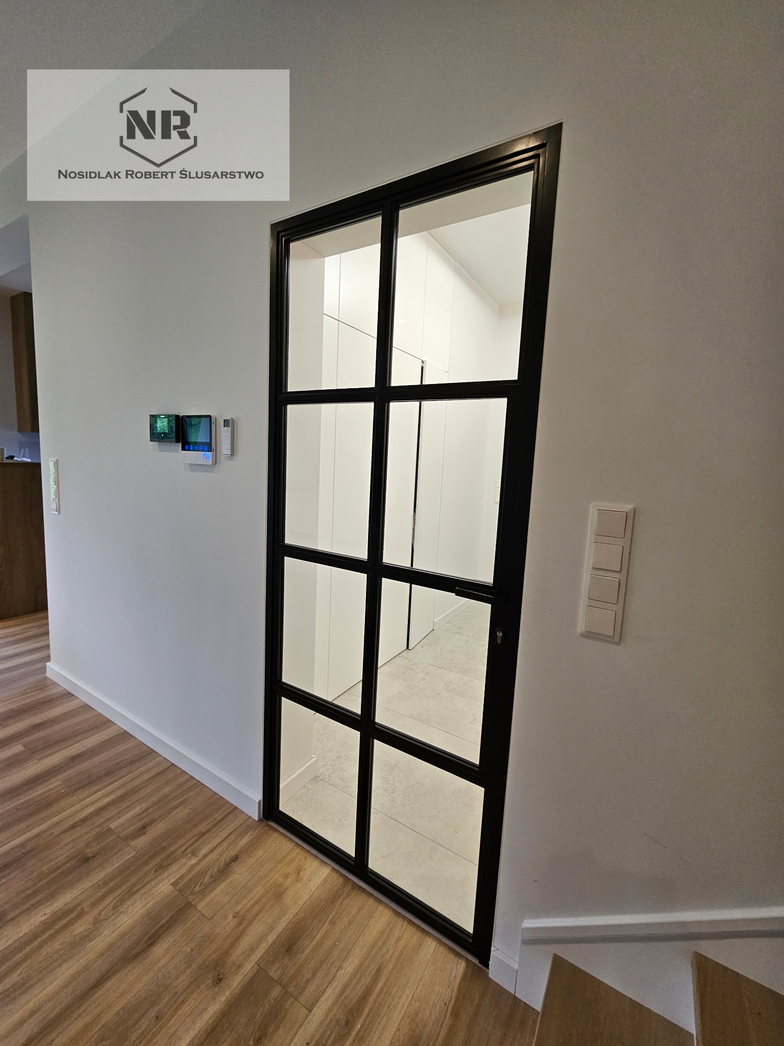 Wewnętrzna przeszklona ścianka działowa z czarnymi szprosami, widok z korytarza z drewnianą podłogą i schodami, na ścianie logo firmy ślusarskiej.