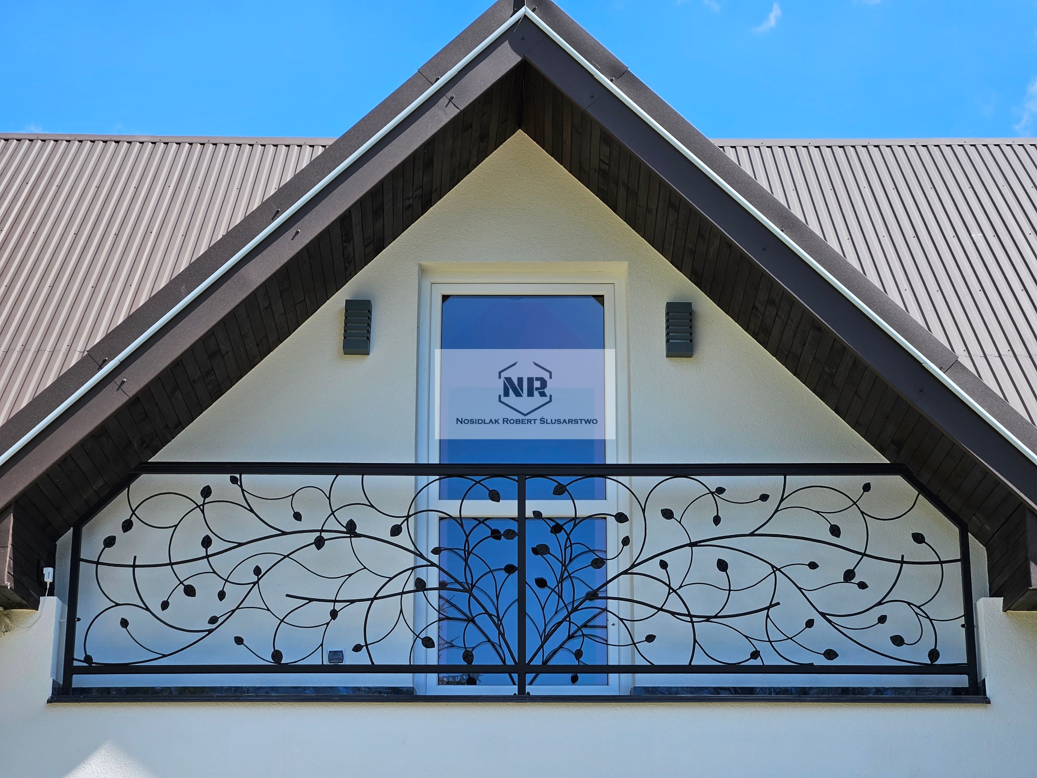 Elegancka, czarna balustrada balkonowa z motywem roślinnym na tle białej ściany domu z oknem i szarą blachodachówką.