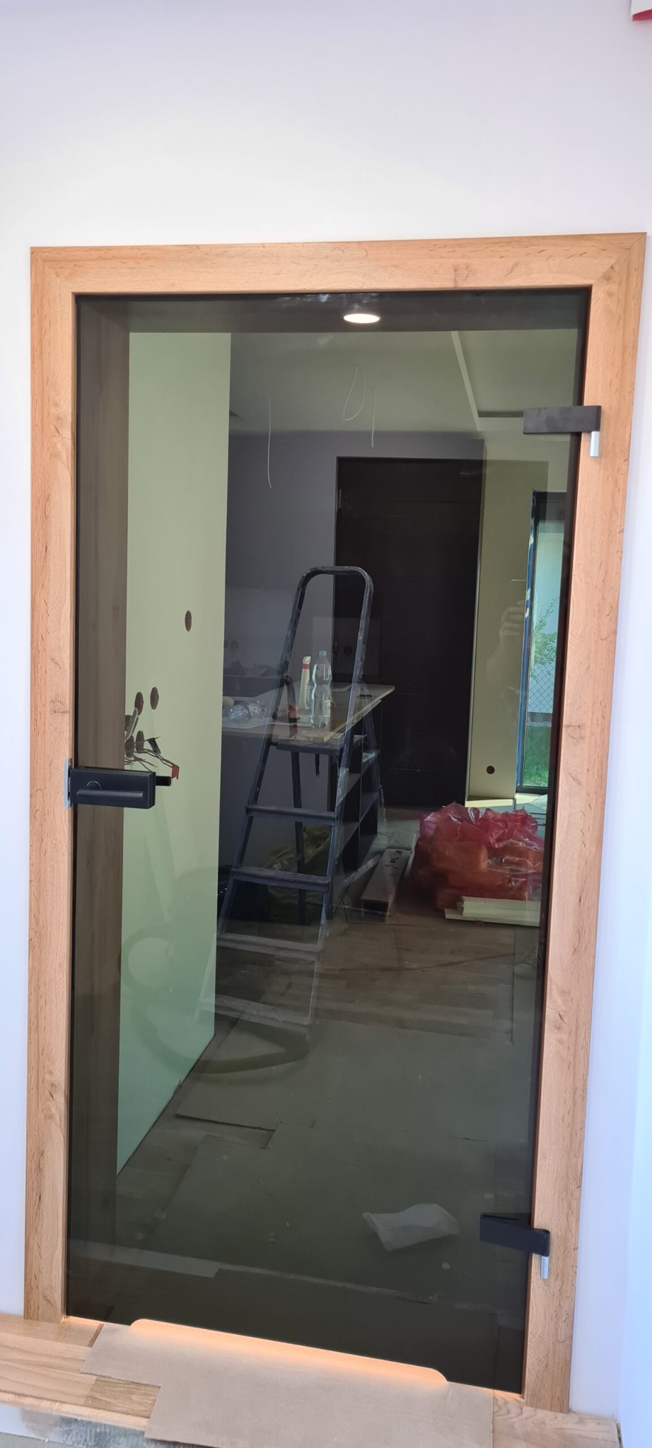 Szklane drzwi z drewnianą ramą, widoczne elementy montażowe, w tle drabina, częściowo wykończone wnętrze.