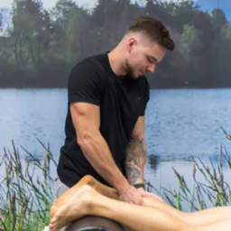 Mężczyzna z tatuażem na ramieniu wykonuje masaż łydek pacjentowi leżącemu na stole z podparciem, w tle widoczna ściana z fototapetą przedstawiającą jezioro i las, w lewym górnym rogu logo Centrum...