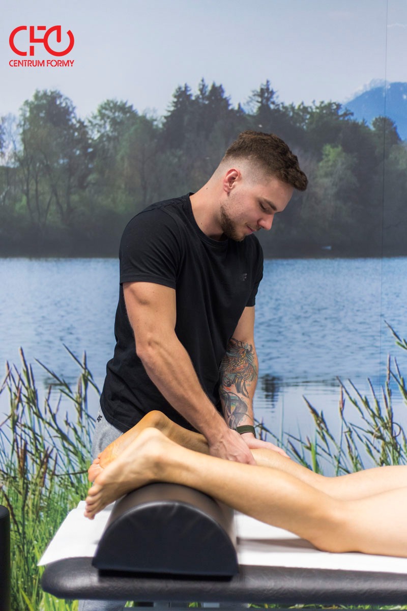 Mężczyzna z tatuażem na ramieniu wykonuje masaż łydek pacjentowi leżącemu na stole z podparciem, w tle widoczna ściana z fototapetą przedstawiającą jezioro i las, w lewym górnym rogu logo Centrum...