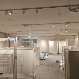Energo Krzysztof Szpakowski - Montaż oświetlenia w nowym sklepie: widoczne reflektory szynowe na suficie, metalowe regały i pracownik na drabinie. Jasne wnętrze w trakcie instalacji.