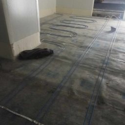 Energo Krzysztof Szpakowski - Rozłożona instalacja ogrzewania podłogowego elektrycznego na folii izolacyjnej w pomieszczeniu przed wylaniem posadzki. Widoczne kable grzewcze i listwy mocujące.