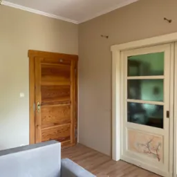 Wnętrze pokoju z dwiema odmiennymi parami drzwi: drewniane drzwi z widoczną strukturą drewna i białe drzwi z szybami i ręcznie malowanym motywem kwiatowym u dołu, na podłodze panele.