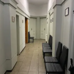 Korytarz biurowy z rzędem czarnych krzeseł, płytkami podłogowymi w odcieniach beżu i ścianami w kolorze jasnej zieleni. W tle widoczne drzwi i grzejnik.