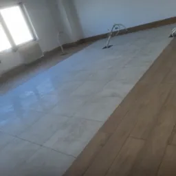 Ukończona podłoga: szare płytki ułożone w karo i panele drewnopodobne, widoczne wystające kable elektryczne z podłogi, jasne wnętrze z oknem.