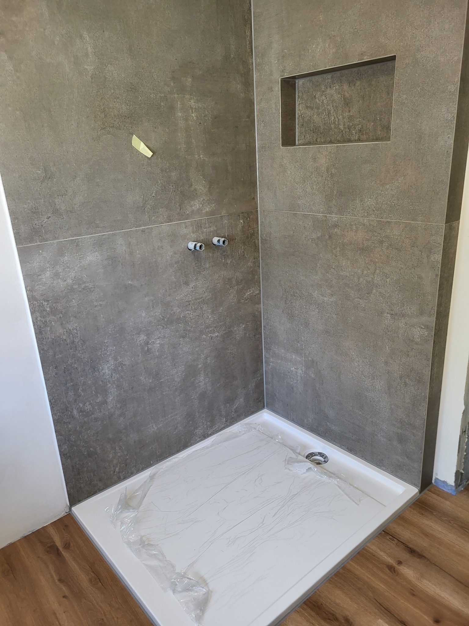 Płytki imitujące beton w kabinie prysznicowej z wnęką, brodzik zabezpieczony folią, widoczne przyłącza wodne i fragment podłogi z paneli.