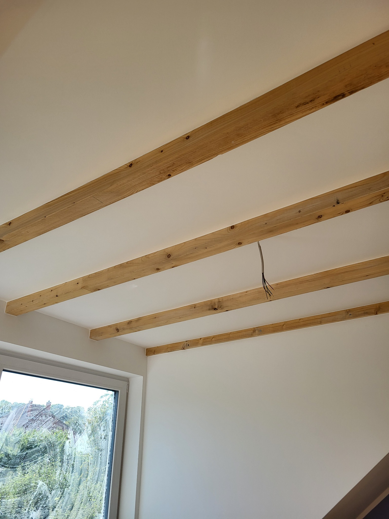 Biały sufit z dekoracyjnymi, drewnianymi belkami, widoczne wystające kable elektryczne i fragment okna z zamazaną szybą.