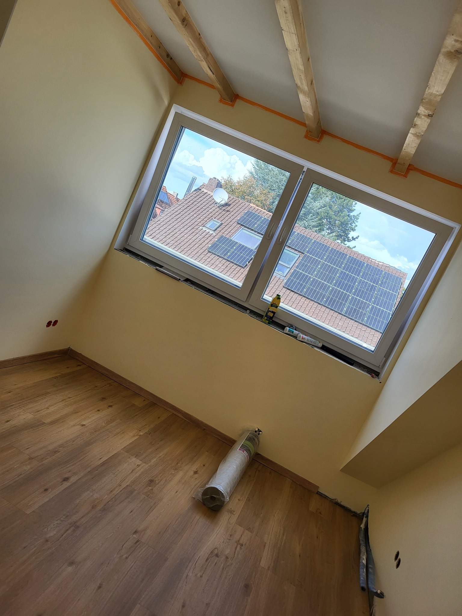 Pokój na poddaszu po remoncie z drewnianą podłogą, świeżo pomalowanymi ścianami w odcieniach beżu oraz drewnianymi belkami stropowymi. Przez okno widać dach z panelami słonecznymi.