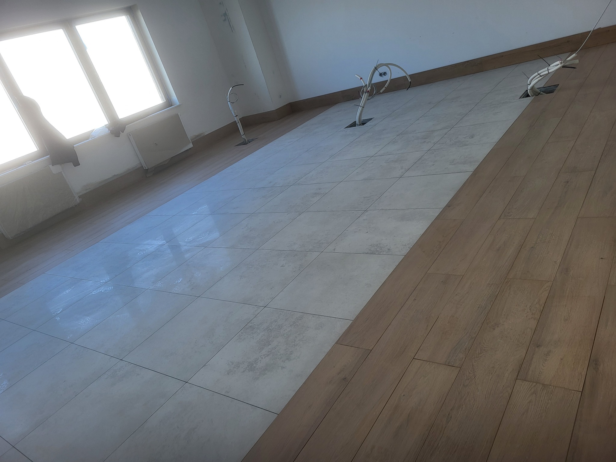 Ukończona podłoga: szare płytki ułożone w karo i panele drewnopodobne, widoczne wystające kable elektryczne z podłogi, jasne wnętrze z oknem.