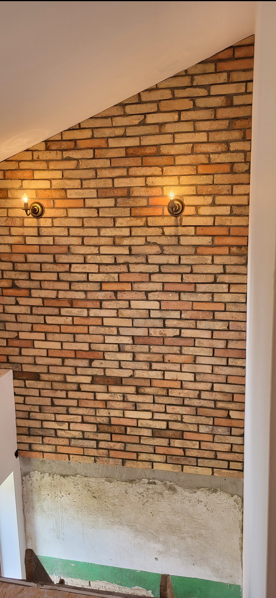 Ściana z cegły klinkierowej w trakcie wykańczania, widoczne dwa kinkiety z żarówkami Edison, dolna część ściany surowa, przygotowana do dalszych prac.