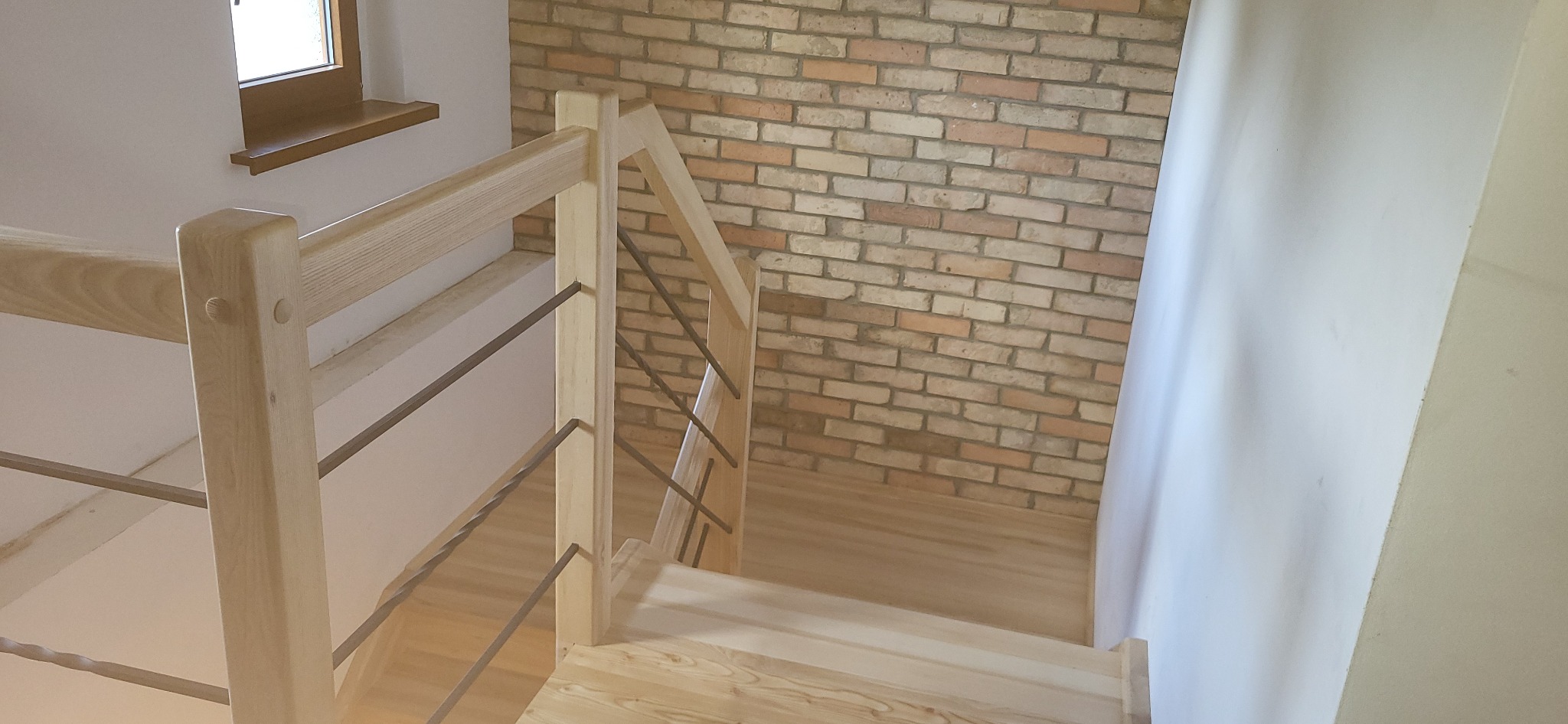 Jasne drewniane schody z metalowymi balustradami, widok z góry na ceglaną ścianę i białe ściany w tle.