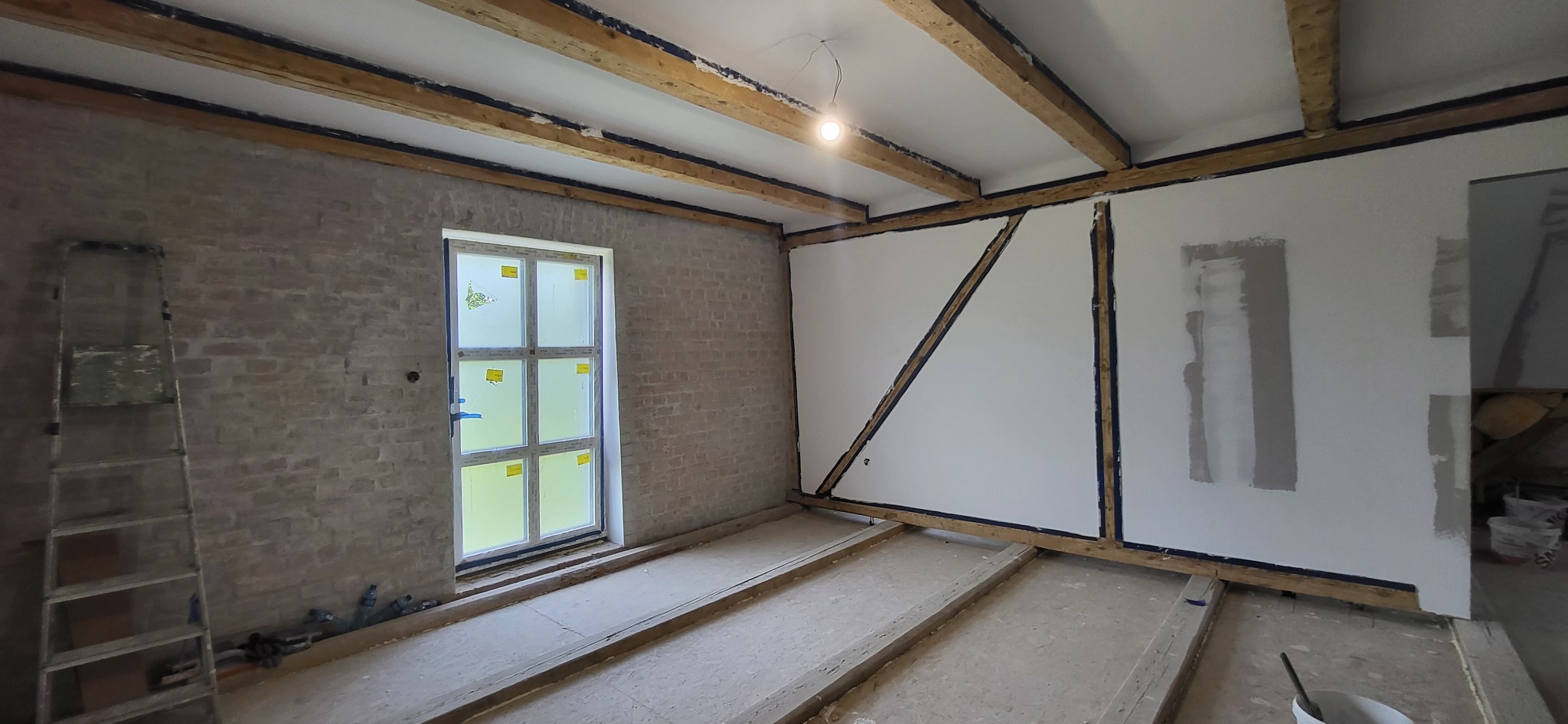 Wnętrze remontowanego pomieszczenia z odsłoniętymi belkami stropowymi, częściowo otynkowaną ceglaną ścianą, przygotowaną do malowania ścianą działową z drewnianą konstrukcją wzmacniającą...