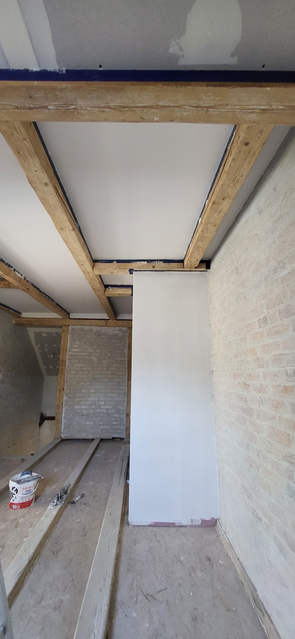 Wnętrze w trakcie prac: sufit z drewnianymi belkami i płytami gipsowymi, częściowo pomalowana ściana z cegły, widoczne narzędzia i wiadro z farbą.